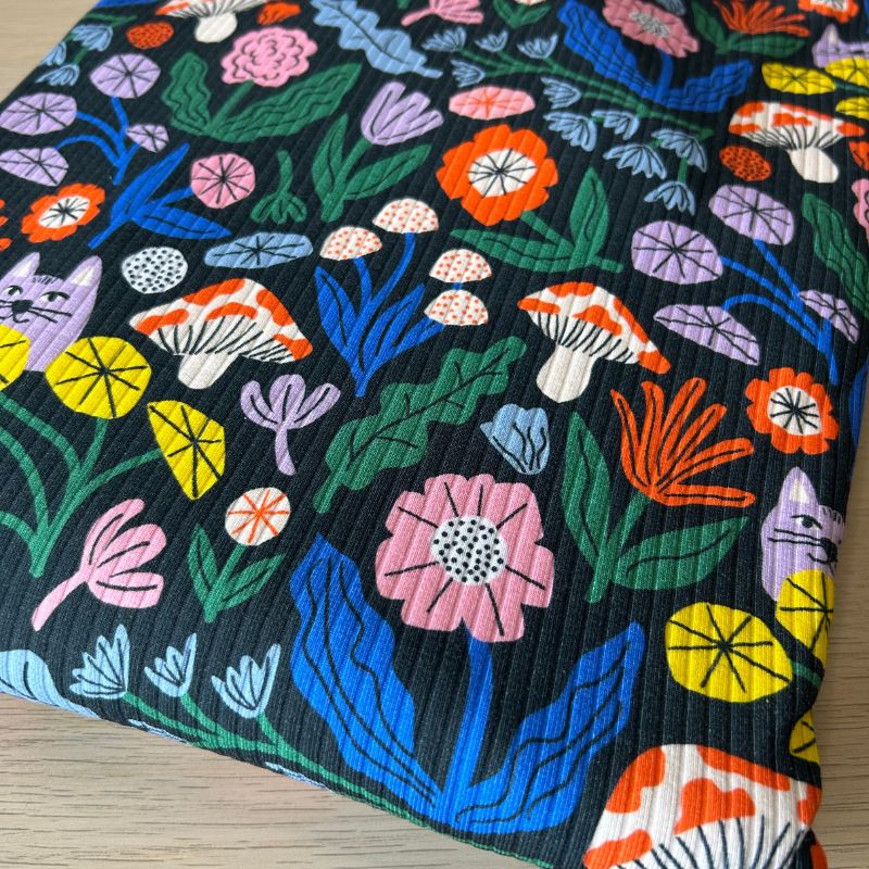 A close-up of a knitted fabric featuring a vibrant, multicolored folk-art style floral and mushroom pattern on a black background, laid flat on a light wood surface.