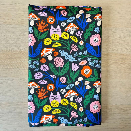 A rectangular fabric sample with a colorful folk-art floral and animal print on a black background, laid flat on a light wood surface.
