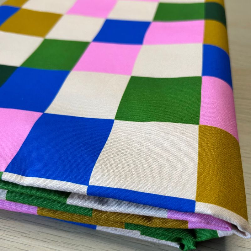 A folded piece of fabric with a colorful patchwork checkered pattern of pink, blue, green, cream, and mustard yellow squares, displayed on a light wood surface.