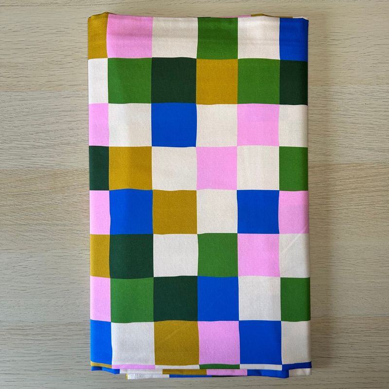A piece of folded fabric  featuring a colorful checkered pattern of squares in pink, blue, green, mustard yellow, and off-white, laid flat on a light wood surface.