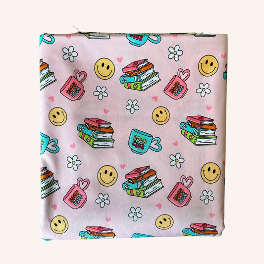 A square fabric sample or napkin featuring a cute school-themed pattern on a light pink background. The pattern includes yellow smiley faces, white daisies, pink hearts, stacks of books, a pink heart-shaped purse, a teal mug, and small pink hearts.