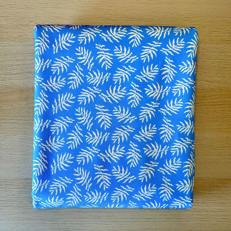 A square fabric item, likely a napkin or small scarf, featuring a white palm leaf pattern on a bright blue background, laid flat on a light wood surface.