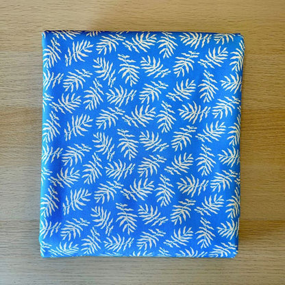 A square fabric item, likely a napkin or small scarf, featuring a white palm leaf pattern on a bright blue background, laid flat on a light wood surface.