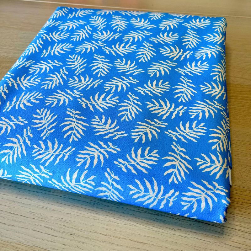 A folded piece of blue fabric with a repeating white palm leaf pattern, displayed on a light wood surface.