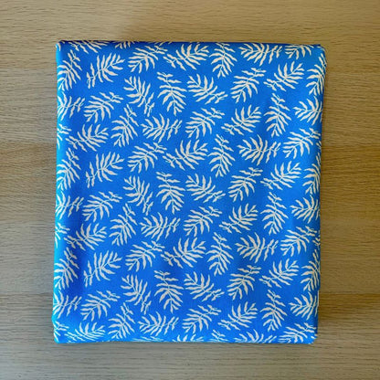 A square fabric item, likely a napkin or small cloth square, featuring a white palm leaf pattern on a bright blue background, laid flat on a light wood surface.
