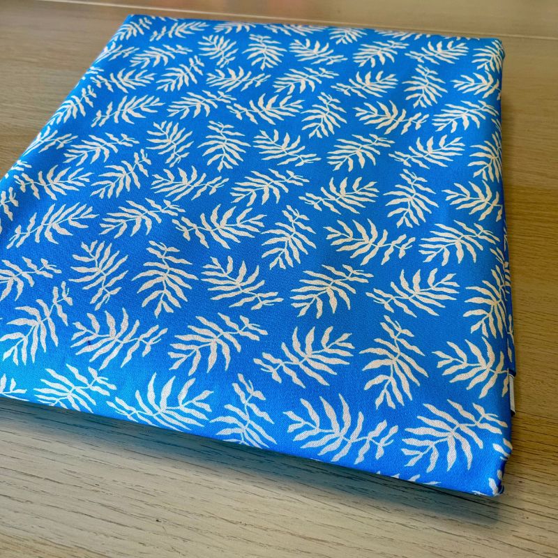 A folded piece of blue fabric with a repeating white palm leaf block print, displayed on a light wood surface.