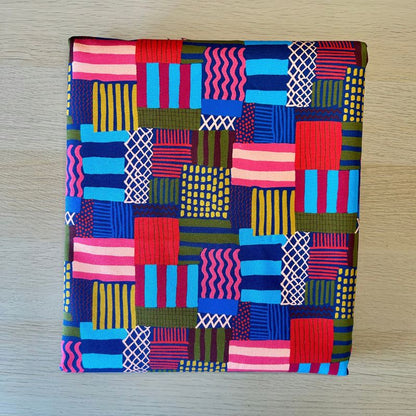 A square fabric pouch or small bag with a colorful geometric patchwork print, laid flat on a light wood surface.