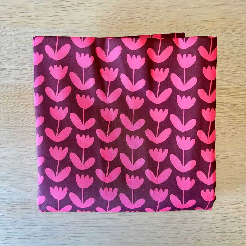 A square fabric item, likely a napkin or small pocket square, featuring a repeating tulip pattern in shades of pink and magenta on a dark plum-purple background, displayed flat on a light wood surface.