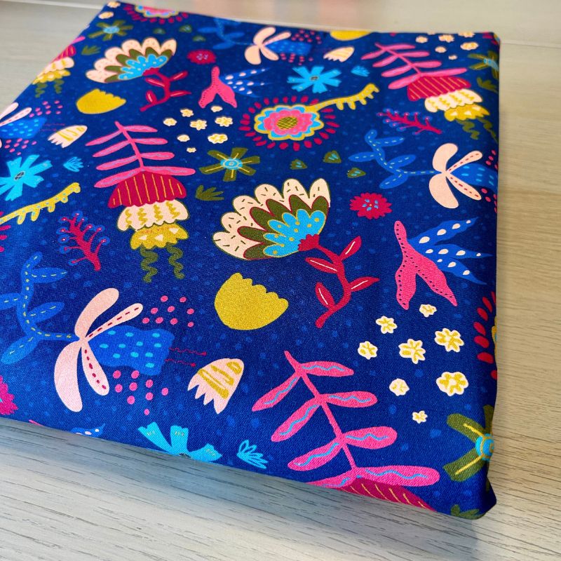A folded piece of fabric with a vibrant, multicolored abstract floral and insect pattern on a deep blue background, displayed on a light wood surface.