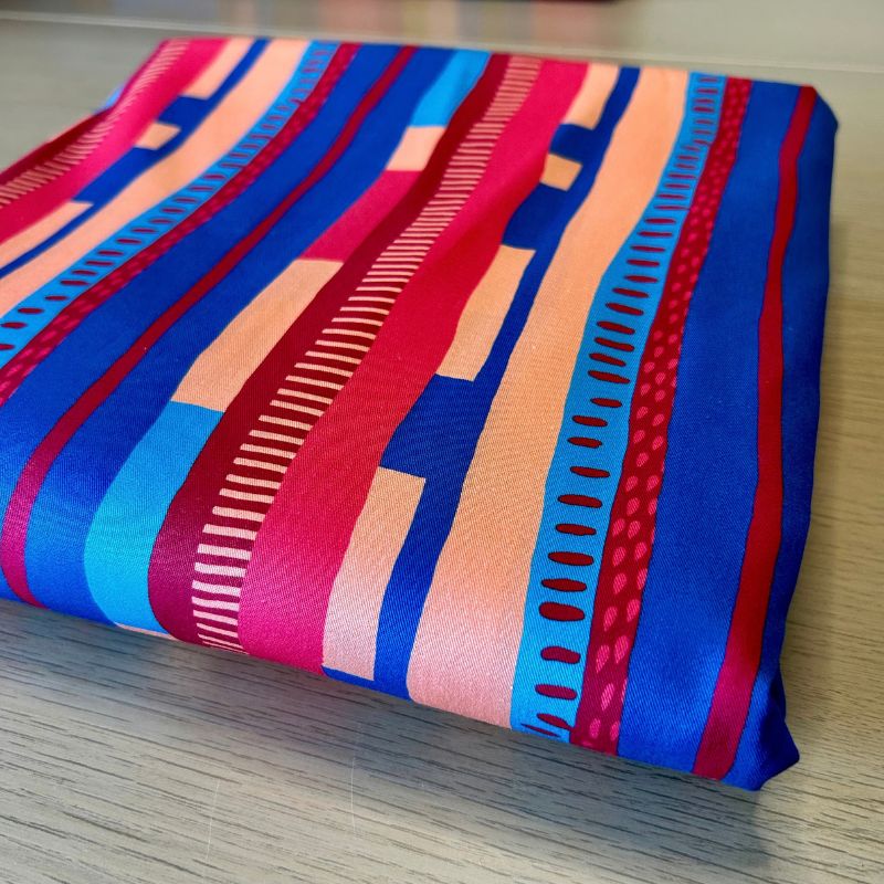 A folded piece of fabric with a vibrant geometric pattern of vertical stripes in blue, red, pink, and peach, displayed on a light wood surface.