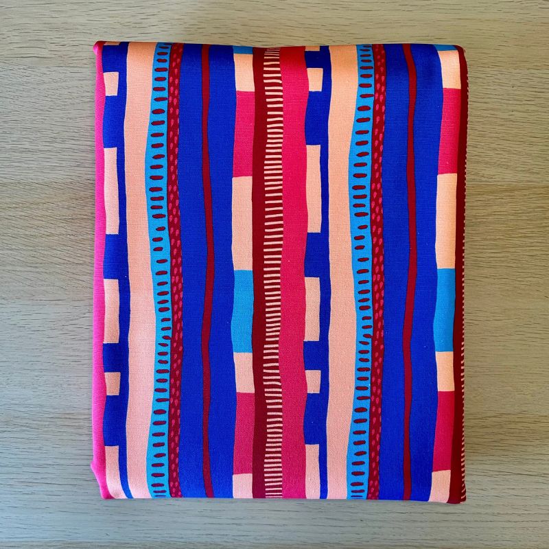 A folded square fabric item, likely a napkin or small scarf, featuring a vibrant geometric pattern with vertical stripes in blue, pink, red, orange, and light blue on a light wood background.