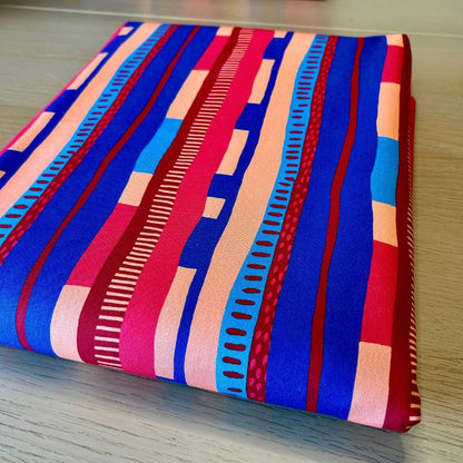 A folded piece of fabric with a bold geometric stripe pattern in blue, red, pink, and light peach, displayed on a light wood surface.