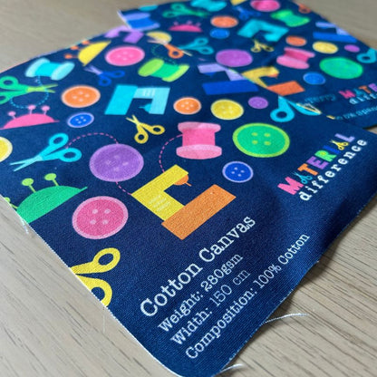 Fabric sample with colorful sewing-themed design and 'Cotton Canvas' label on a wooden surface.