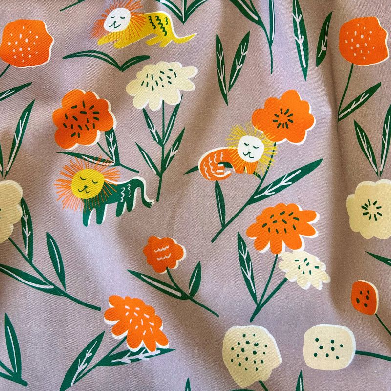 A close-up of a fabric featuring stylized orange flowers, cream blossoms, green leaves, and small cartoon lions on a dusty lilac background.