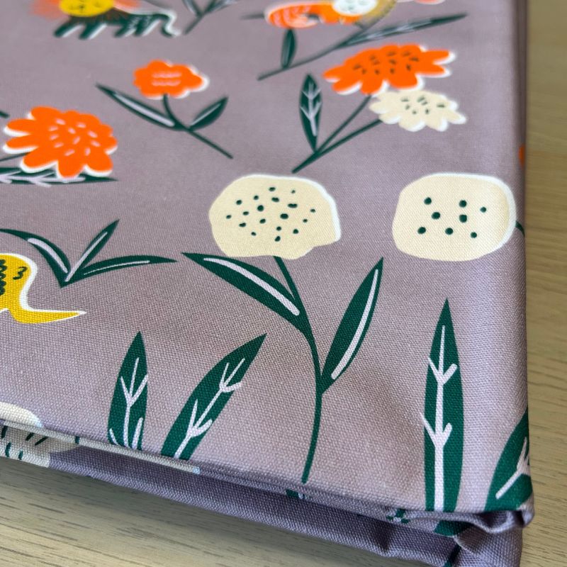 A folded piece of fabric with a muted mauve-purple background and a colorful floral pattern featuring orange, cream, and yellow flowers with dark green leaves, displayed on a light wood surface.