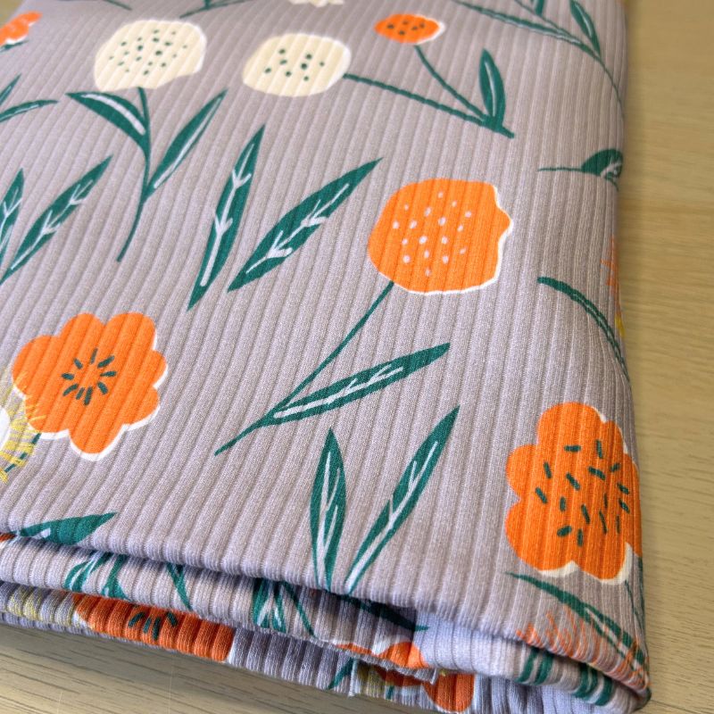 A folded piece of ribbed fabric with a floral pattern in orange, cream, and green on a muted lavender-gray background, placed on a light wood surface.