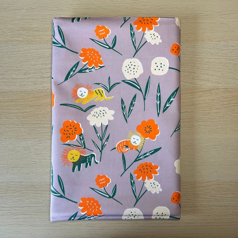 A folded piece of  fabric featuring a whimsical floral and bird pattern on a lilac background, laid flat on a light wood surface.