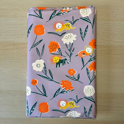 A folder piece of fabric featuring a whimsical pattern of orange flowers, white flowers, yellow lions, and green leaves on a dusty purple background, laid flat on a light wood surface.