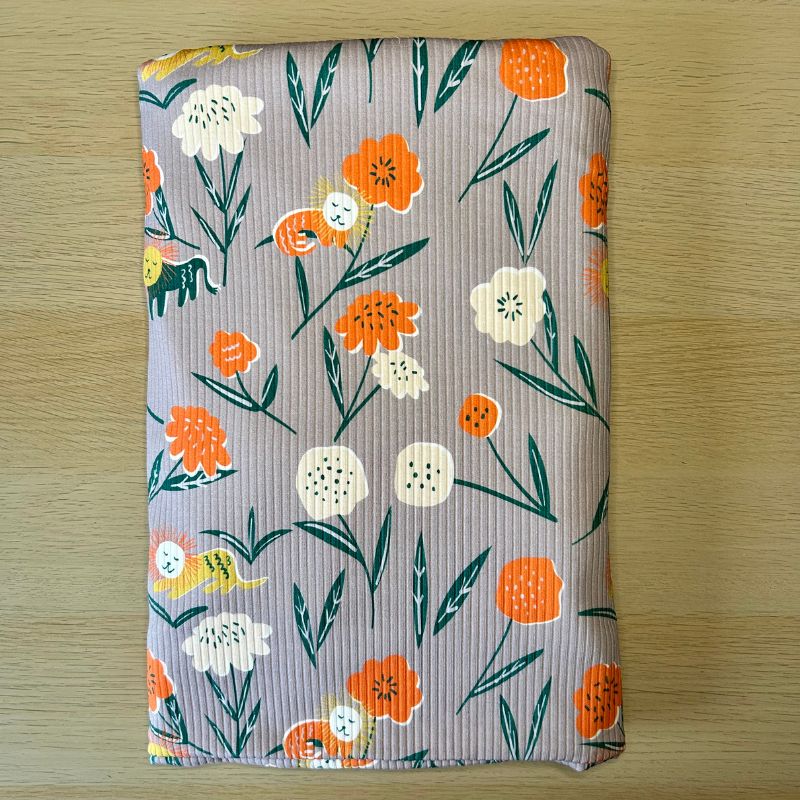 A folded piece of ribbed knit fabric featuring a colorful floral and lion pattern on a dusty purple background, placed on a light wood surface.