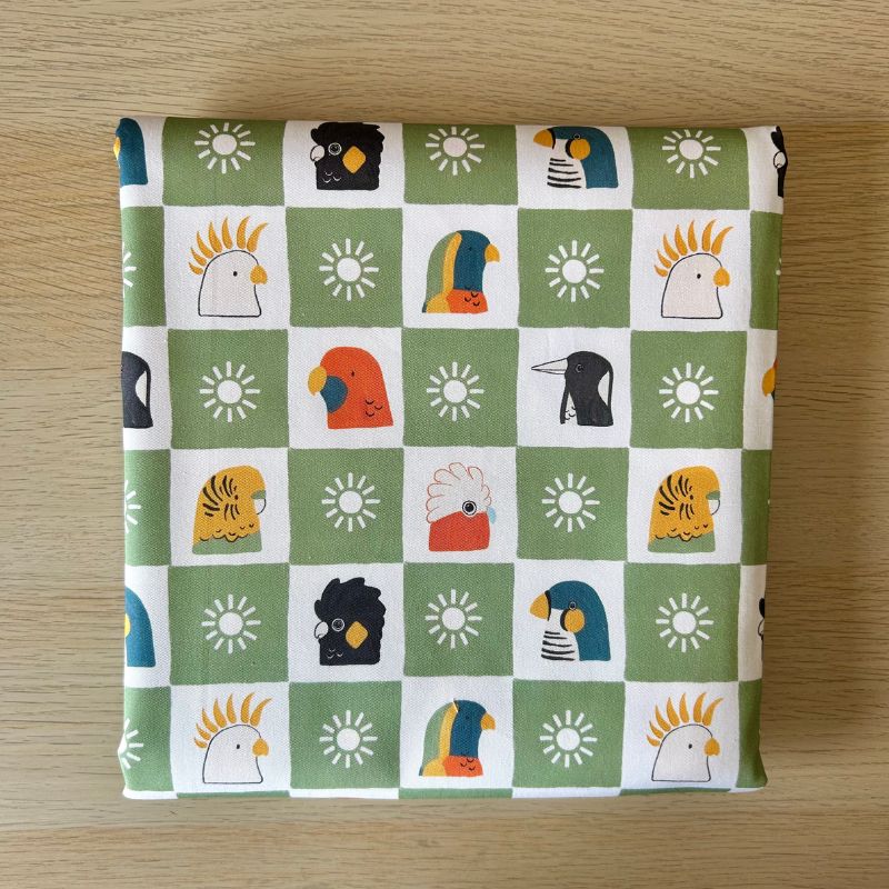 Fabric featuring a green and white checkered pattern with various colorful bird illustrations and sun motifs, laid flat on a light wood surface.