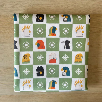 Fabric featuring a green and white checkered pattern with various colorful bird illustrations and sun motifs, laid flat on a light wood surface.