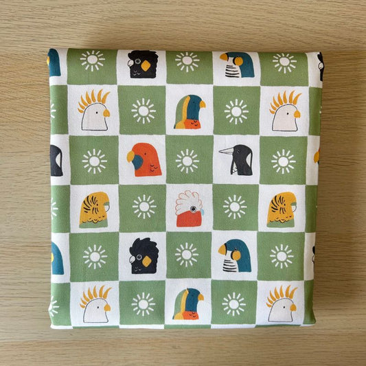 Fabric featuring a green and white checkered pattern with various colorful bird illustrations and sun motifs, laid flat on a light wood surface.