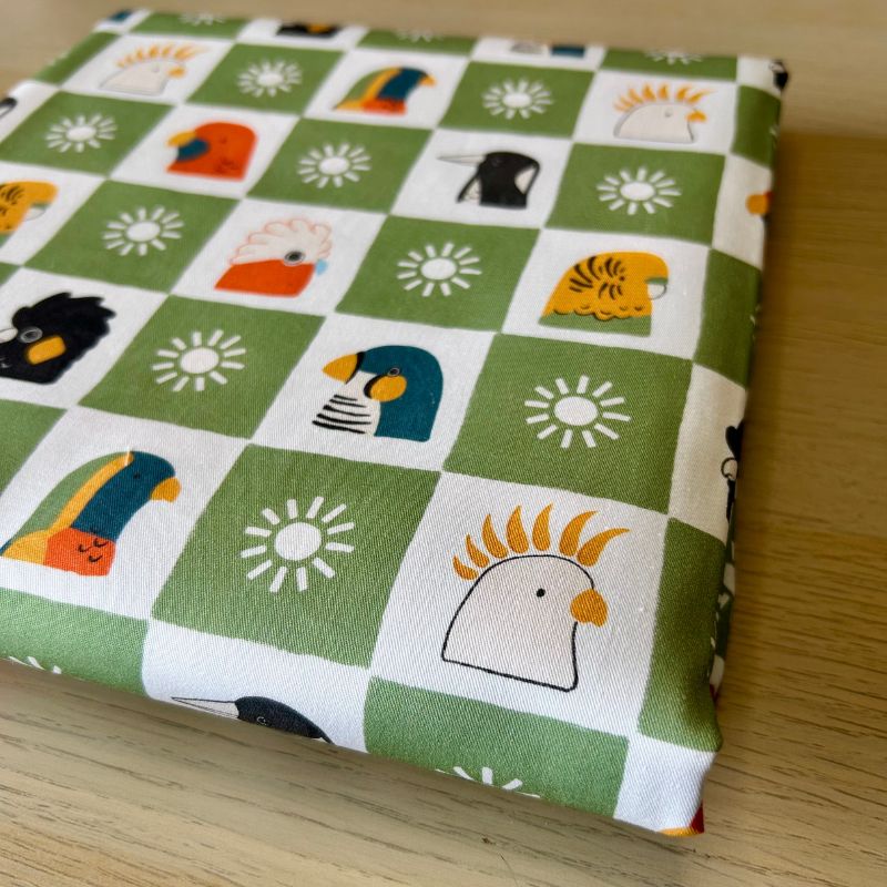 Cotton fabric featuring a green and white checkered pattern with cartoon birds and sun motifs, displayed on a light wood surface.
