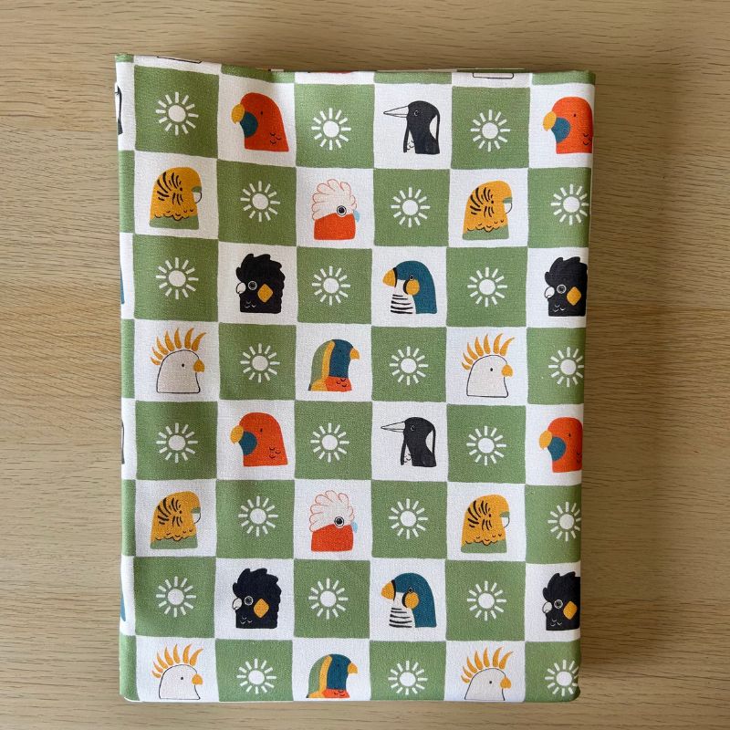 Cotton fabric featuring a green and white checkered pattern with stylized cockatoos and sun motifs, laid flat on a light wood surface.