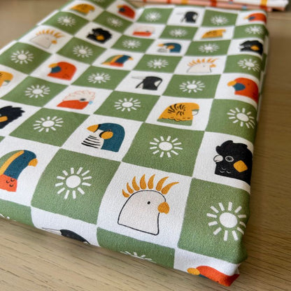 A folded piece of fabric with a green and white checkered pattern featuring cartoon birds, flowers, and suns, displayed on a light wood surface.