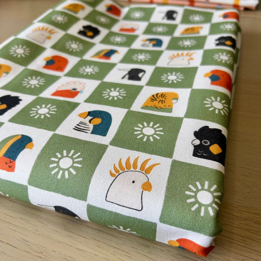A folded piece of fabric with a green and white checkered pattern featuring cartoon birds, flowers, and suns, displayed on a light wood surface.